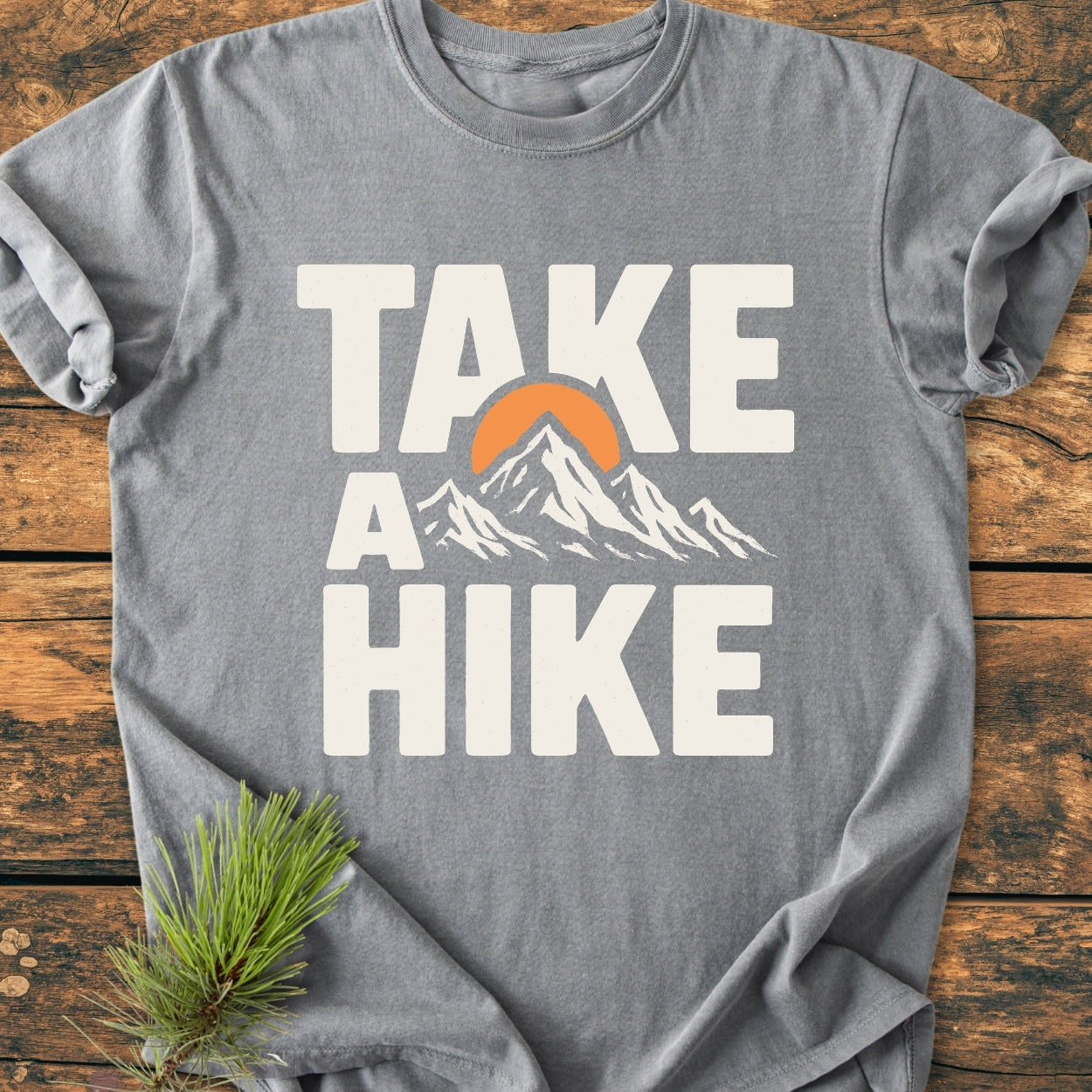 Take A Hike - Vintage Wash Tee featuring a white slogan with a mountain and sun graphic. Laid on wood, this gray, soft-washed, 100% cotton t-shirt offers a relaxed, durable fit.
