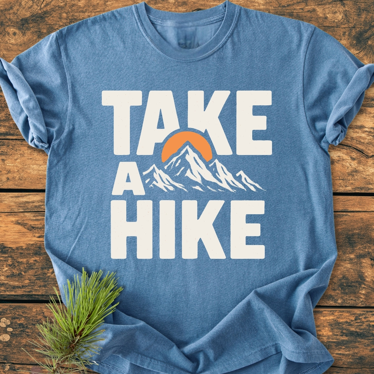 Take A Hike - Vintage Wash Tee featuring a heather-blue color with a bold “TAKE A HIKE” print, stylized mountain range, and orange sun. Laid on wooden surface with a pine branch beside it.