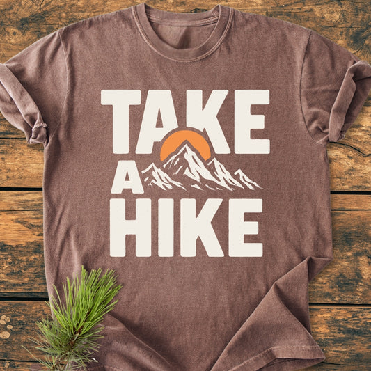 Take A Hike - Vintage Wash Tee: Brown short-sleeved T-shirt with TAKE A HIKE and mountain graphic, laid on wood. Pine sprig adds rustic touch, emphasizing its outdoor theme and relaxed fit.