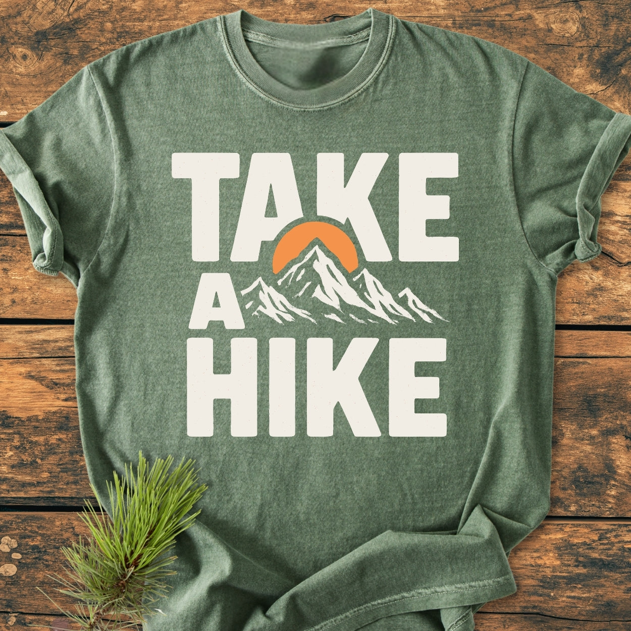 Take A Hike - Vintage Wash Tee: Green T-shirt with “TAKE A HIKE” text, mountain range, and setting sun graphic; laid on wood with a pine sprig, highlighting its outdoorsy design.