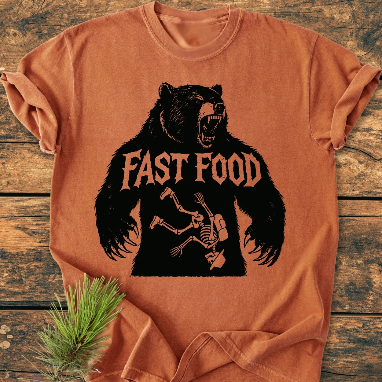 Fast Food - Vintage Wash Tee features a roaring bear silhouette with FAST FOOD text, humorously displaying a skeleton inside. Laid flat on wood, pine needles add rustic charm.