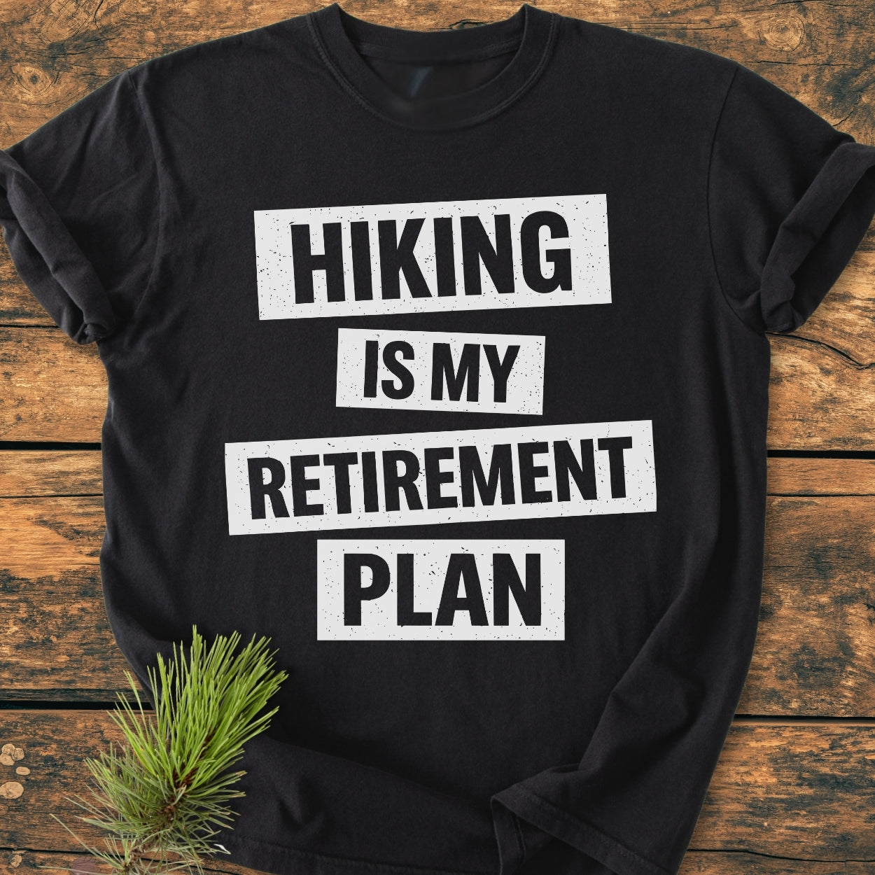 The Hiking Is My Retirement Plan - Vintage Wash Tee features bold white text on a black, soft-washed, garment-dyed tee, styled with a pine sprig on a wooden surface.