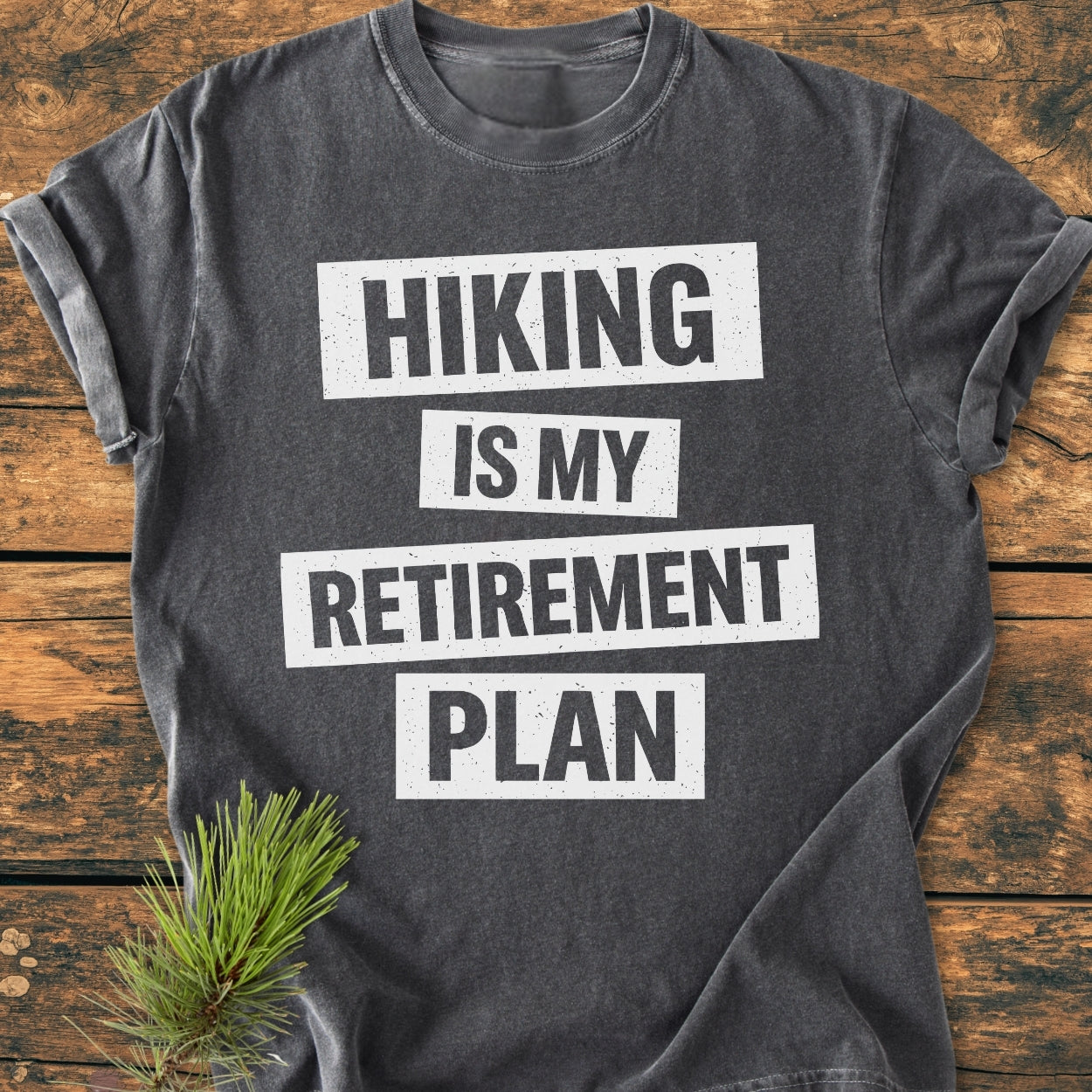 Charcoal-gray Hiking Is My Retirement Plan - Vintage Wash Tee with bold white text, laid flat on wood. Features durable, soft-washed, 100% ring-spun cotton. Pine sprig accents the rustic theme.