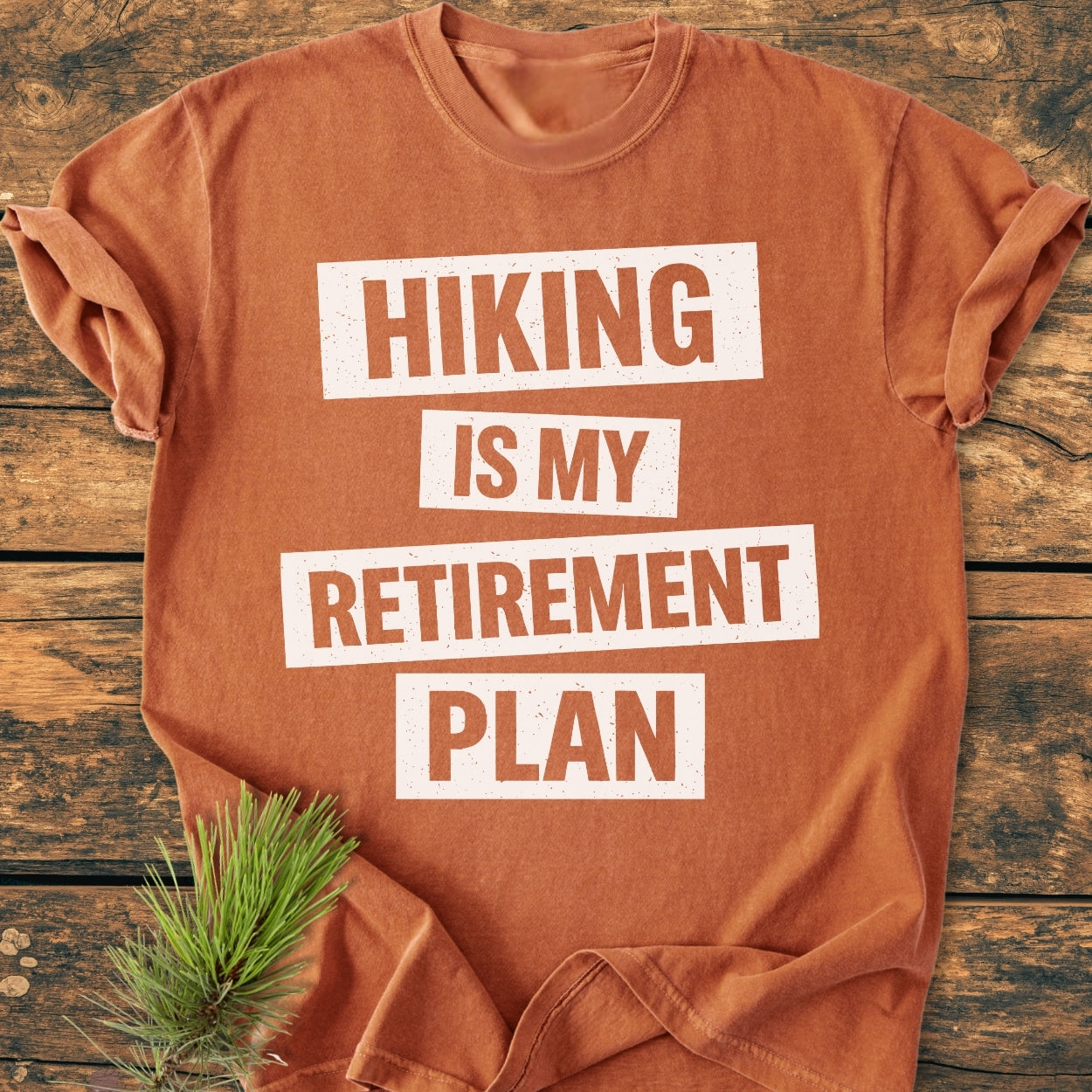 Hiking Is My Retirement Plan - Vintage Wash Tee displayed with rolled sleeves, featuring bold white print on a rust-orange base. Accented by a pine sprig on a wooden surface.