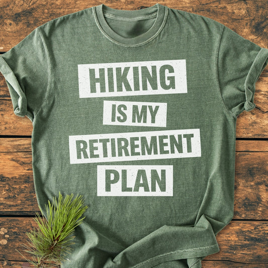 Green crew-neck tee, Hiking Is My Retirement Plan in white block letters, with a rustic pine accent. Made of soft, durable, 100% ring-spun cotton for a relaxed fit.