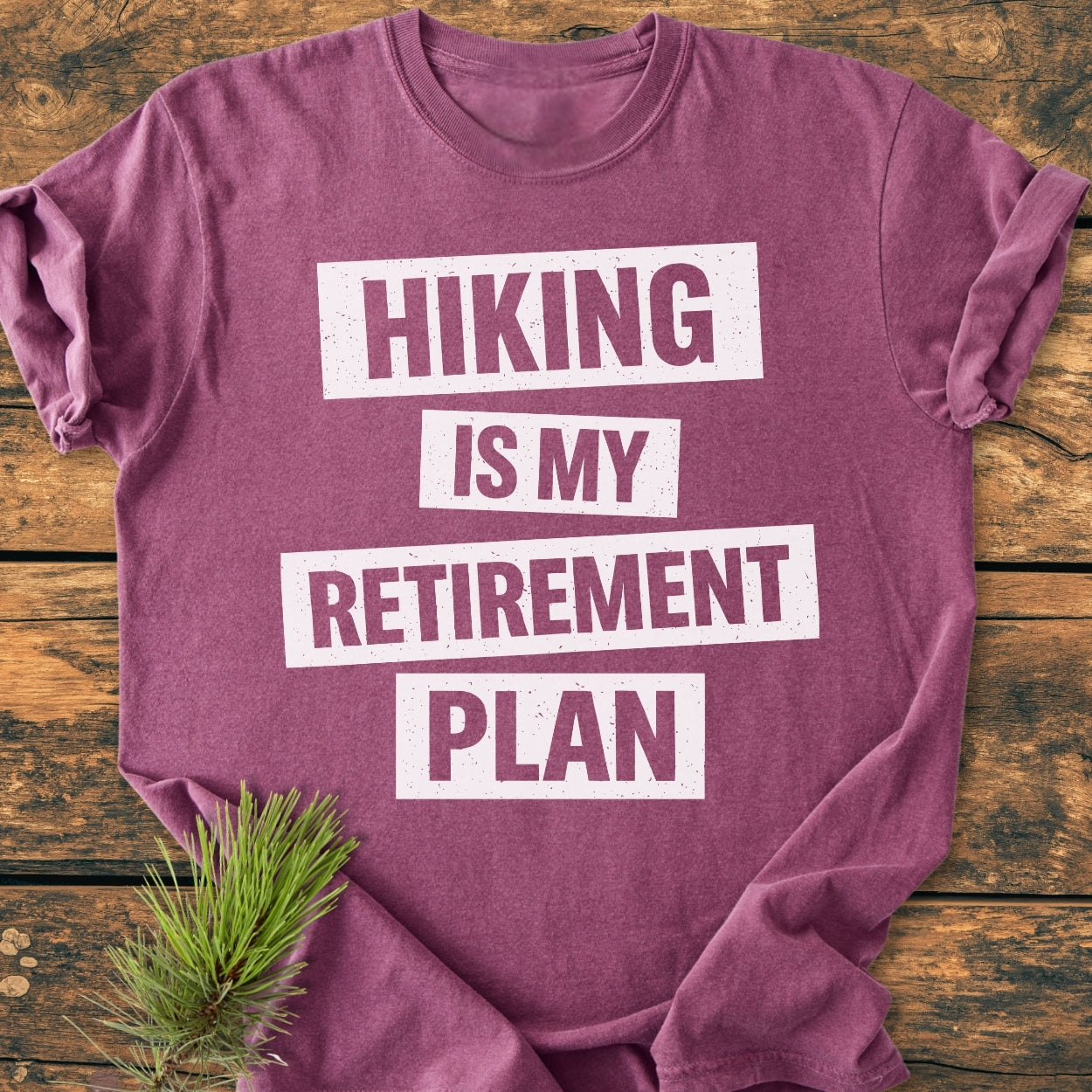 Laid-flat, heathered plum tee reads HIKING IS MY RETIREMENT PLAN in white. Sprig of pine nearby, showcasing the Hiking Is My Retirement Plan - Vintage Wash Tee on a wooden surface.