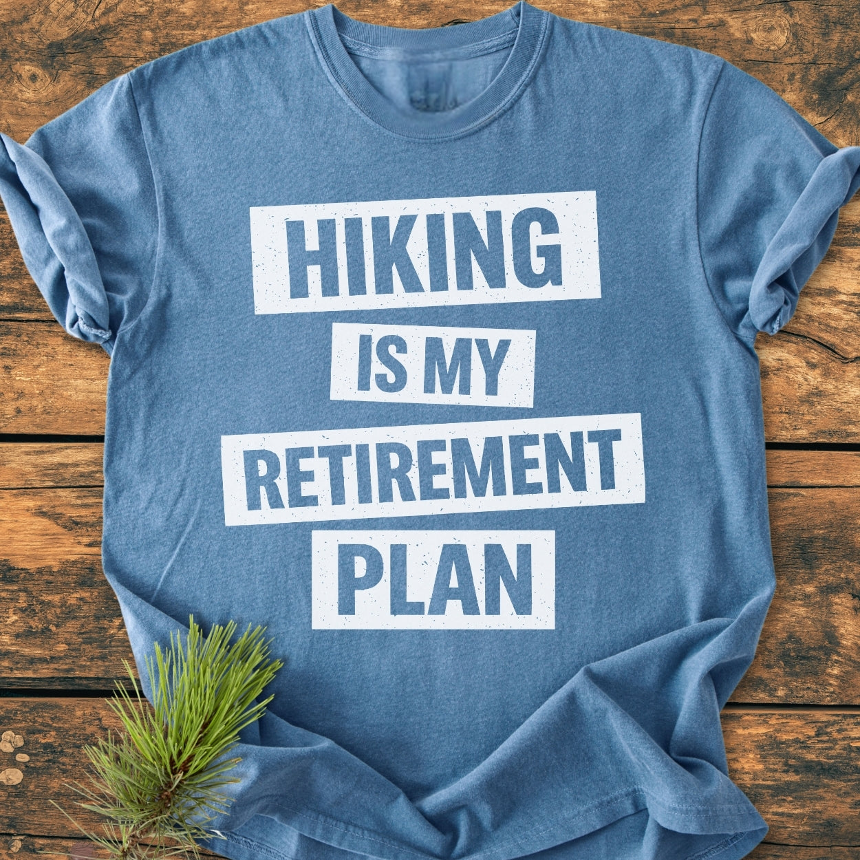 Hiking Is My Retirement Plan - Vintage Wash Tee, heather-blue, with bold white text. Laid flat on wood, paired with a pine sprig, emphasizing its outdoorsy theme and relaxed fit.