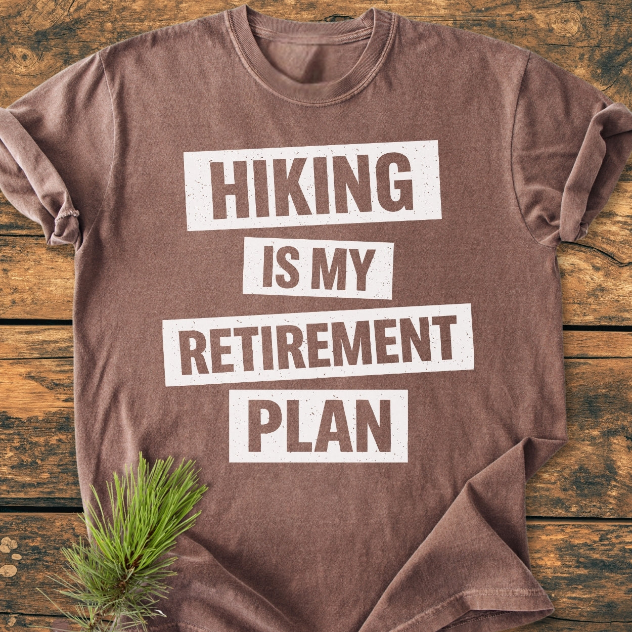 Brown Hiking Is My Retirement Plan - Vintage Wash Tee made of soft-washed, garment-dyed cotton with bold white lettering, laid flat with an evergreen sprig beside it on a wooden surface.