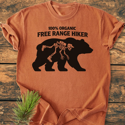 Organic Free Range Hiker - Vintage Wash Tee displaying a bear graphic with a hiker skeleton and slogan, laid on wood with a pine sprig, emphasizing an outdoorsy, rustic vibe.