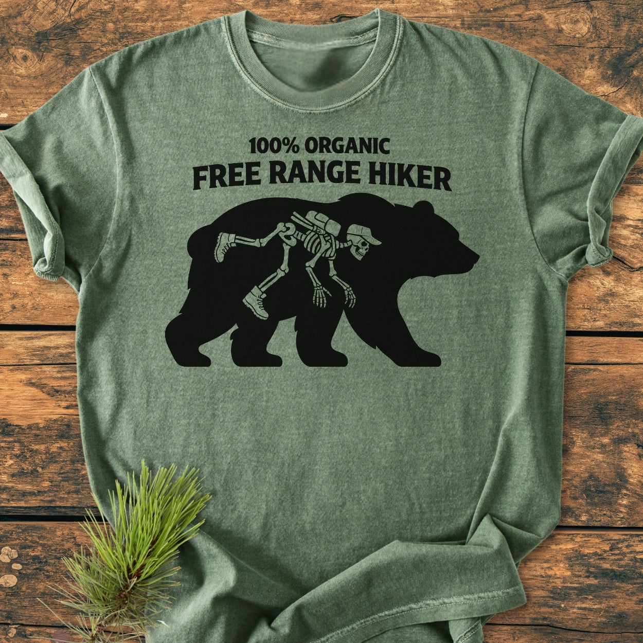 Organic Free Range Hiker - Vintage Wash Tee features a black bear silhouette with a skeletal hiker, text above, on a green T-shirt laid on a wooden surface, highlighting its cozy, durable design.