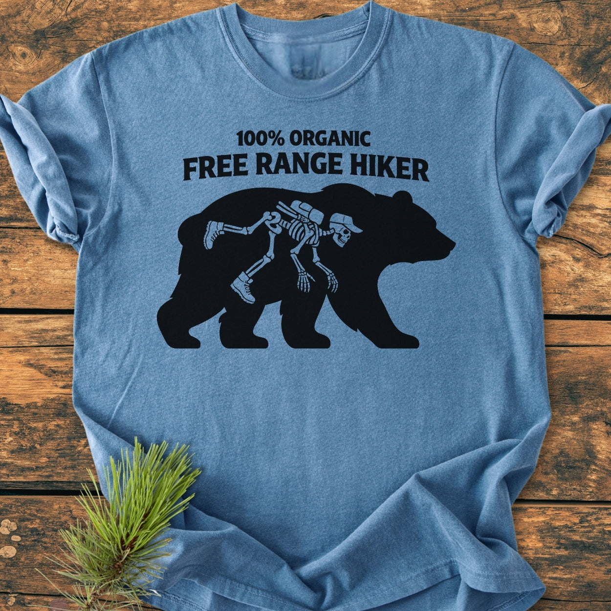 Organic Free Range Hiker - Vintage Wash Tee: Heather-blue T-shirt displaying a humorous design with “100% ORGANIC FREE RANGE HIKER” text and a bear silhouette containing a cartoon hiker skeleton, laid on a wooden surface.