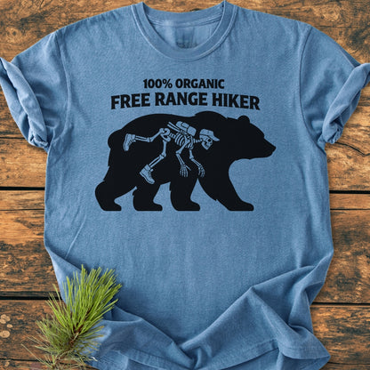 Organic Free Range Hiker - Vintage Wash Tee: Heather-blue T-shirt displaying a humorous design with “100% ORGANIC FREE RANGE HIKER” text and a bear silhouette containing a cartoon hiker skeleton, laid on a wooden surface.