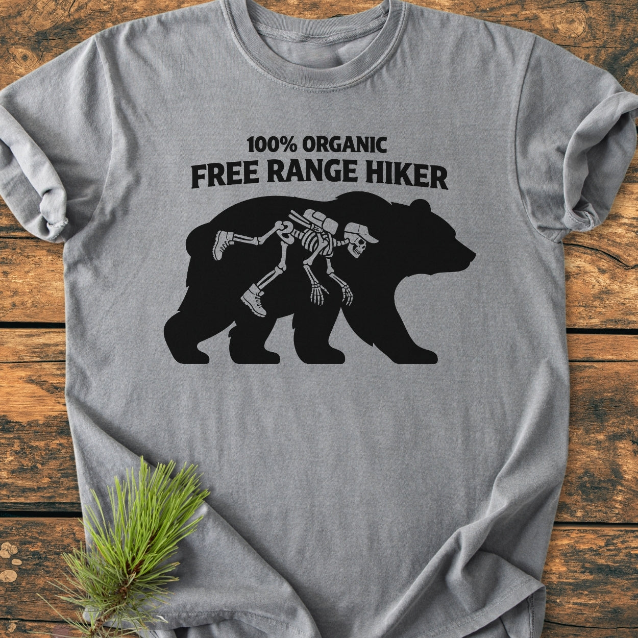 Organic Free Range Hiker - Vintage Wash Tee featuring a humorous bear silhouette with a hiker's skeleton inside. Laid on wood, with pine sprig detail, highlights its casual, cozy design.