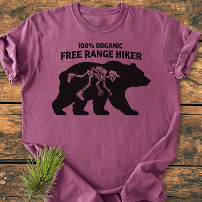 Organic Free Range Hiker - Vintage Wash Tee: Heather-purple T-shirt featuring a whimsical bear design with hiker skeleton, laid on wood with pine sprig; soft, durable, relaxed fit, showcasing Comfort Colors’ garment-dyed quality.