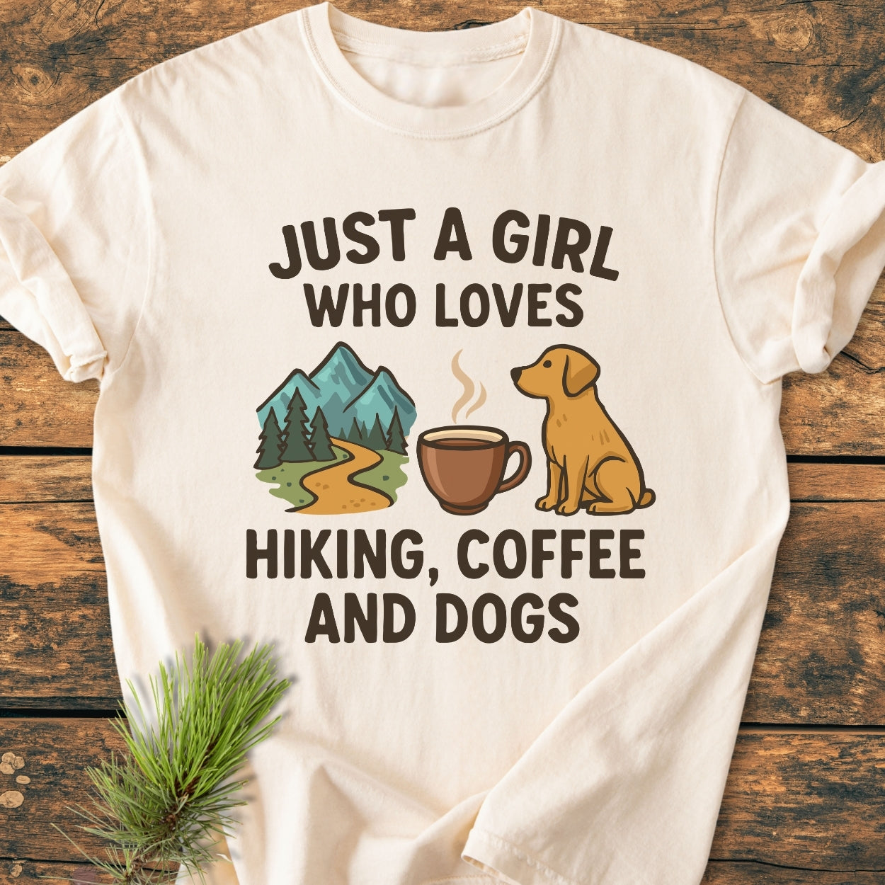 Just A Girl... Hiking Coffee Dogs - Vintage Wash Tee features bold lettering with mountain, coffee, and dog icons, laid on wood, complemented by an evergreen sprig.
