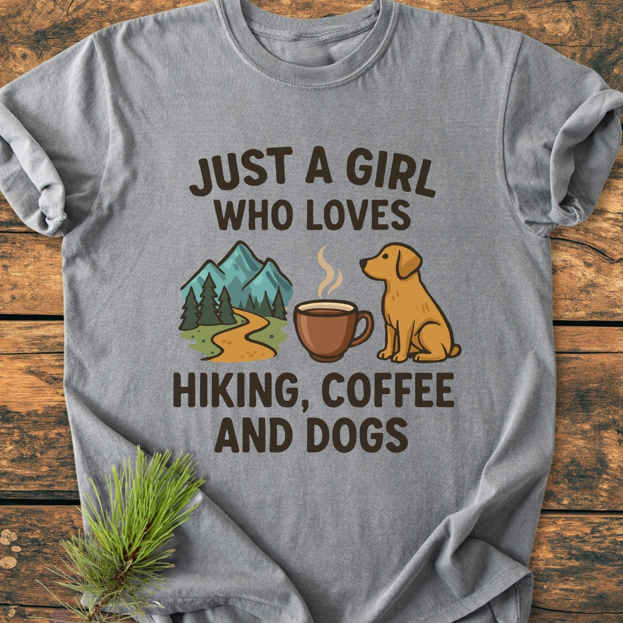 Just A Girl Hiking Coffee Dogs - Vintage Wash Tee features a playful graphic with hiking, coffee, and dog icons on a soft, heather-gray shirt, laid on wood with a pine sprig.