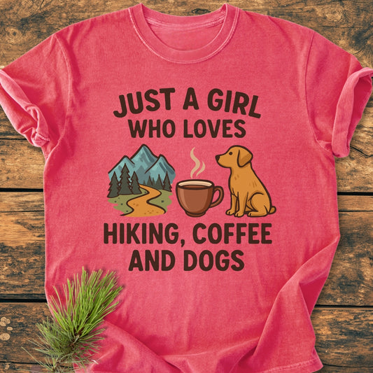 The Just A Girl Who Loves Hiking, Coffee, and Dogs vintage wash tee features whimsical graphics and text on a red T-shirt, displayed on a wooden tabletop with a pine sprig beside it.