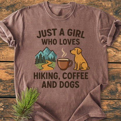 Just A Girl... Hiking Coffee Dogs - Vintage Wash Tee: Brown short-sleeved shirt with hiking, coffee, and dog-themed text and icons, laid flat on a wood surface with a pine sprig.