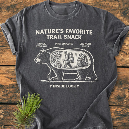 Trail Snack - Vintage Wash Tee features a white bear graphic with labeled compartments on a charcoal-gray shirt, laid on wood. It humorously highlights nature as a snack with “NATURE’S FAVORITE TRAIL SNACK” text.