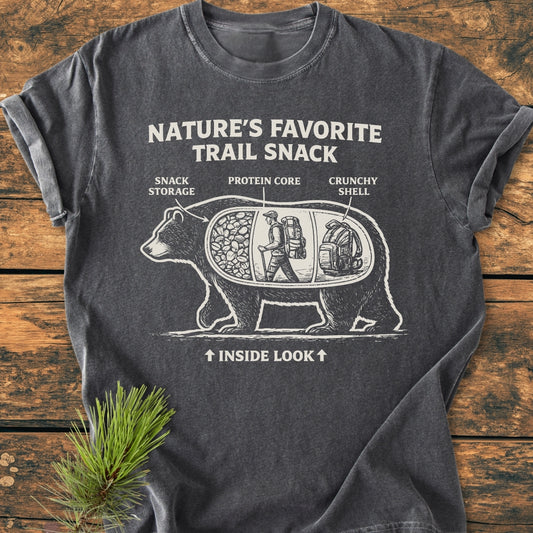 Trail Snack - Vintage Wash Tee features a white bear graphic with labeled compartments on a charcoal-gray shirt, laid on wood. It humorously highlights nature as a snack with “NATURE’S FAVORITE TRAIL SNACK” text.