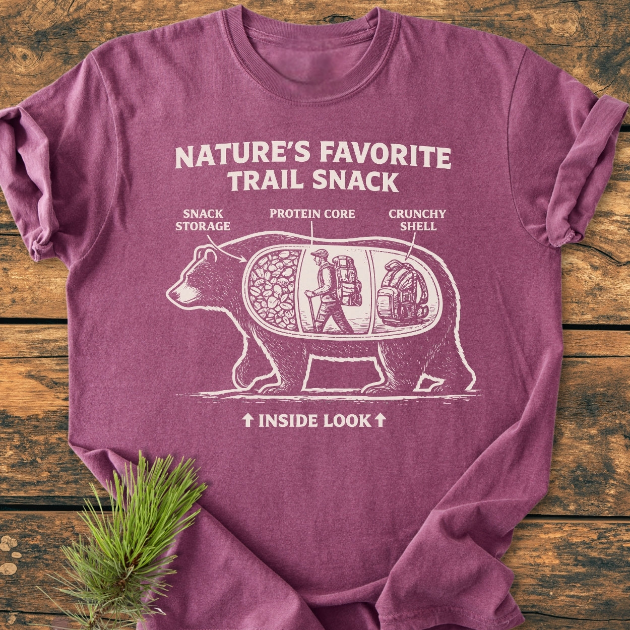 Trail Snack - Vintage Wash Tee featuring a whimsical bear diagram on a maroon-purple shirt, highlighting sections like “Snack Storage” and “Protein Core,” laid on wood with pine sprig.