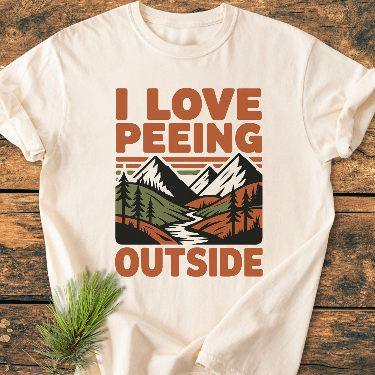 I Love Peeing Outside - Vintage Wash Tee, featuring an earth-toned outdoor scene with pine trees and mountains, laid on a wooden surface, showcasing its relaxed fit and soft-washed, garment-dyed texture.