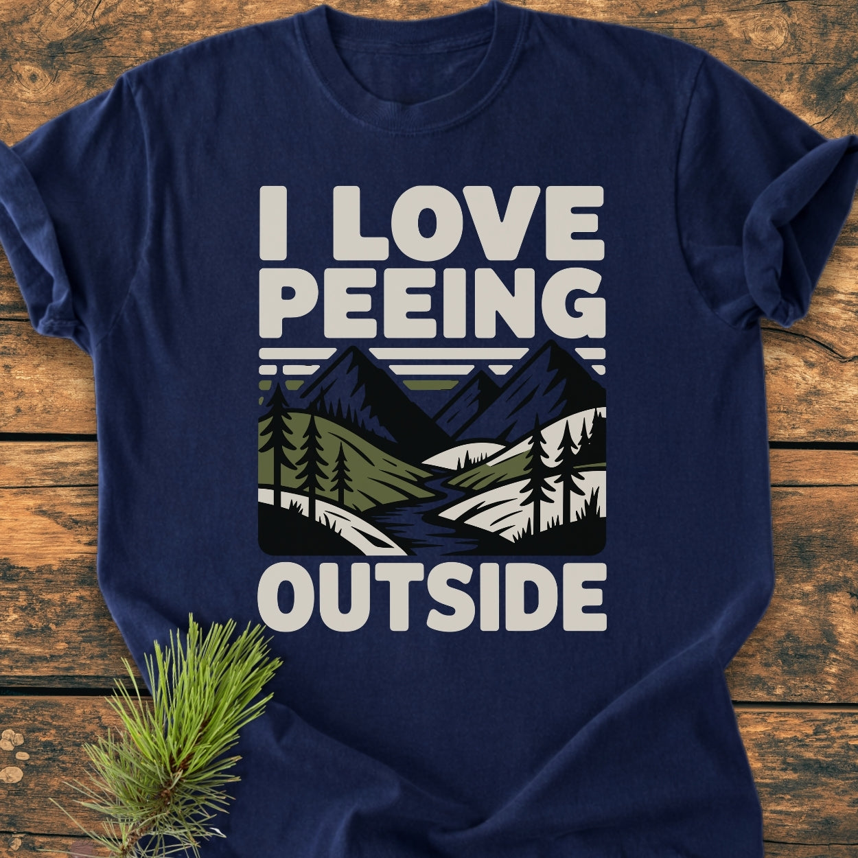 Navy-blue I Love Peeing Outside - Vintage Wash Tee on rustic wood, featuring bold text and nature graphic. Adorned with a pine sprig, illustrating its casual, outdoor-inspired design.