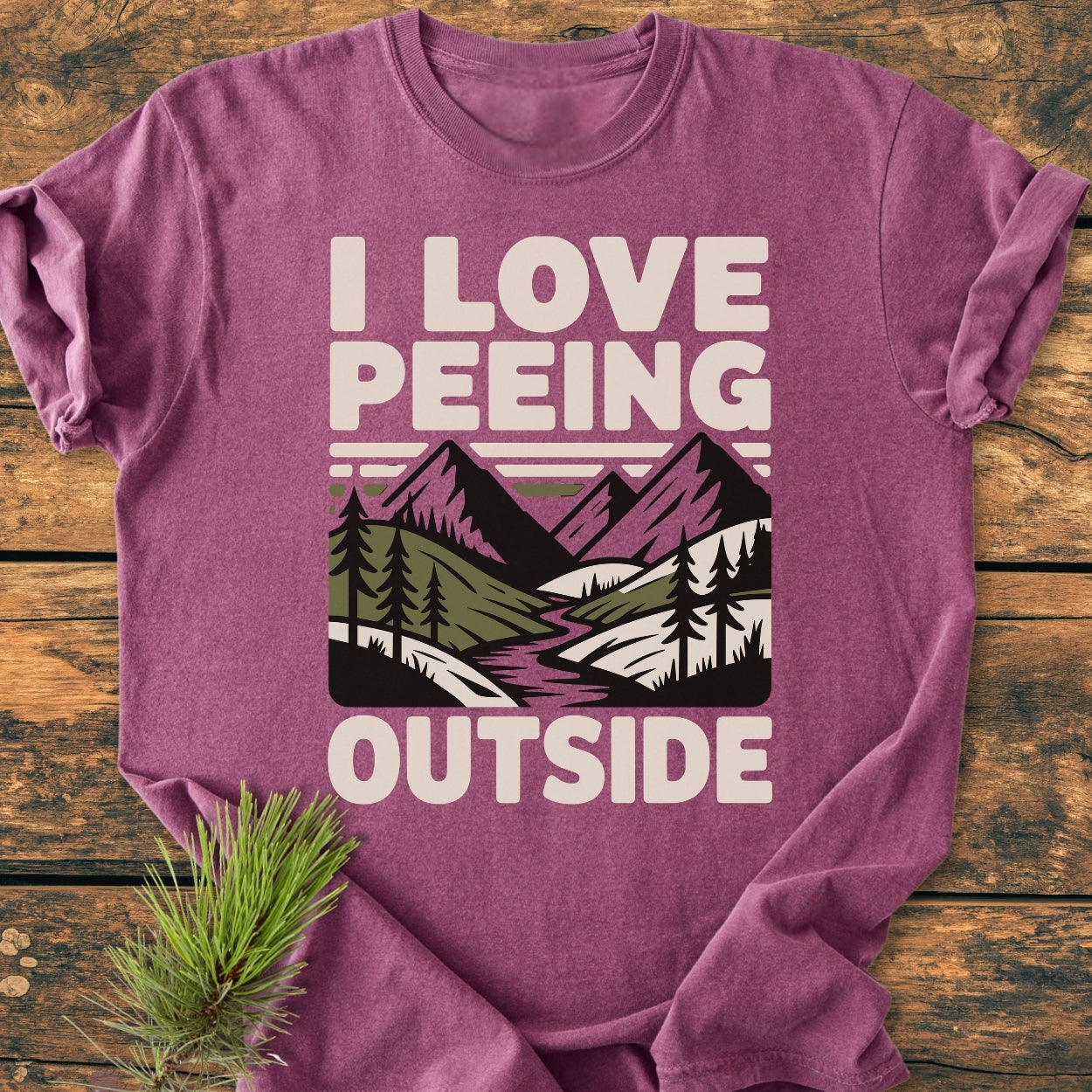 I Love Peeing Outside - Vintage Wash Tee, featuring bold text with a mountain landscape graphic, displayed on wood with a pine sprig. Made from soft, durable, pre-shrunk cotton for comfort.