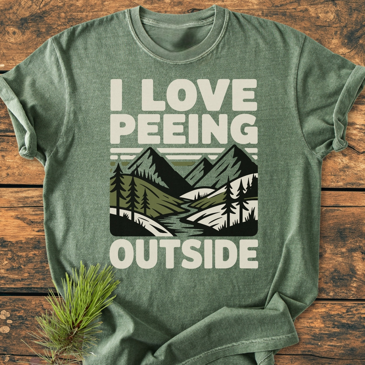 I Love Peeing Outside - Vintage Wash Tee features a mountain-and-pine graphic on heather-green fabric, laid on wood with a pine sprig, embodying an outdoorsy, relaxed vibe.