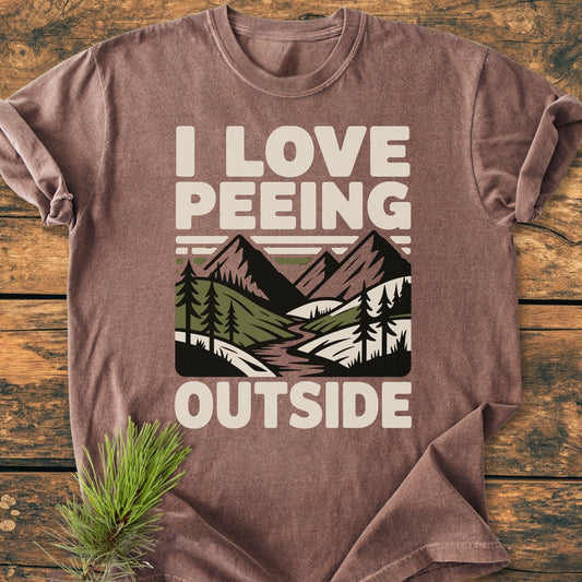 I Love Peeing Outside - Vintage Wash Tee features a stylized outdoor scene with mountains and trees on a soft, brownish-tan shirt, laid on wood with a pine sprig accent.