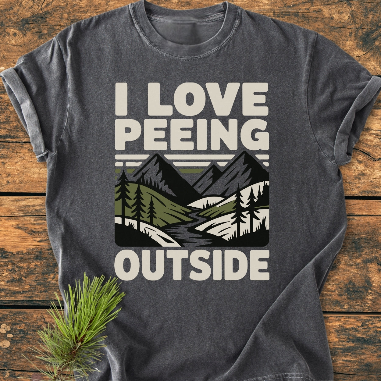 I Love Peeing Outside - Vintage Wash Tee features a mountain-and-river graphic with pine trees on a dark-gray, short-sleeve T-shirt, laid flat with a pine sprig beside it.