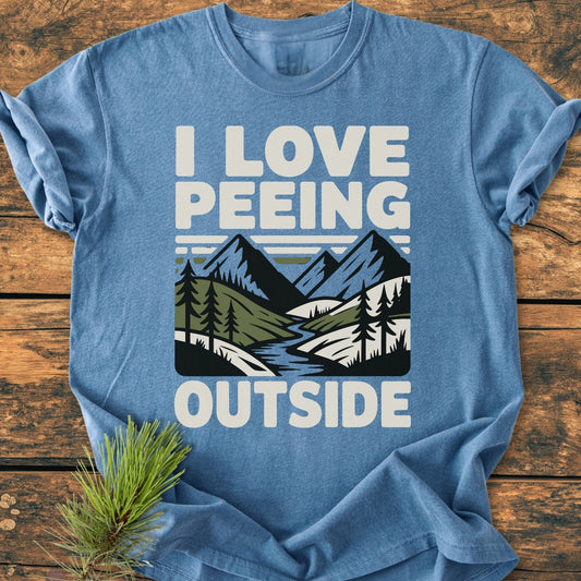 I Love Peeing Outside - Vintage Wash Tee featuring a nature scene with mountains and trees, printed on a heather-blue, soft-washed, durable T-shirt, laid flat on a wooden surface.