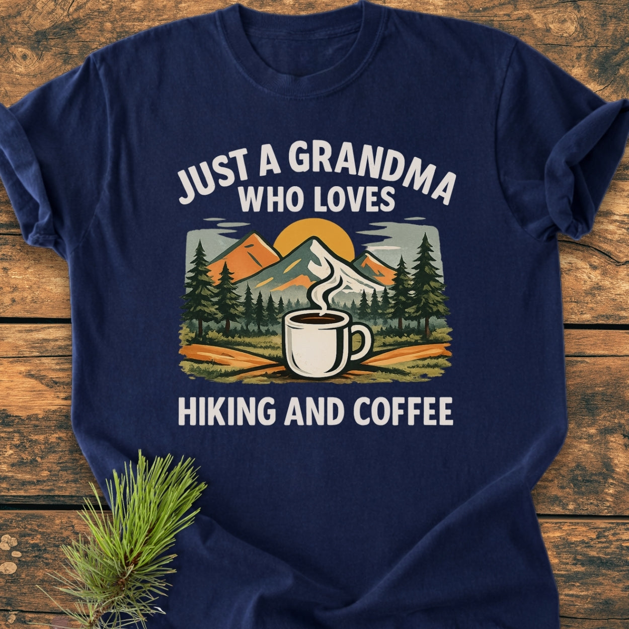 Grandma Loves Hiking And Coffee - Vintage Wash Tee features a rustic graphic of mountains, pine trees, and a coffee mug. Laid on wood, it captures an outdoorsy vibe.