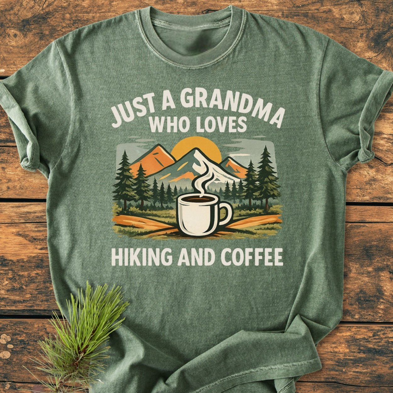 Grandma Loves Hiking And Coffee - Vintage Wash Tee features a stylized outdoor scene with coffee, pine trees, and mountains, printed on a heather-green shirt, laid on a wooden surface.