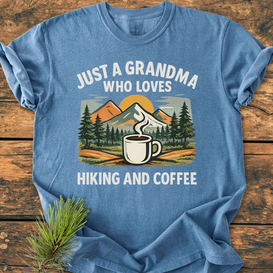 Grandma Loves Hiking And Coffee - Vintage Wash Tee: Heather-blue, folded, with a mountain scene and coffee mug graphic, white text highlights love for hiking and coffee, on a wooden surface with pine sprig.