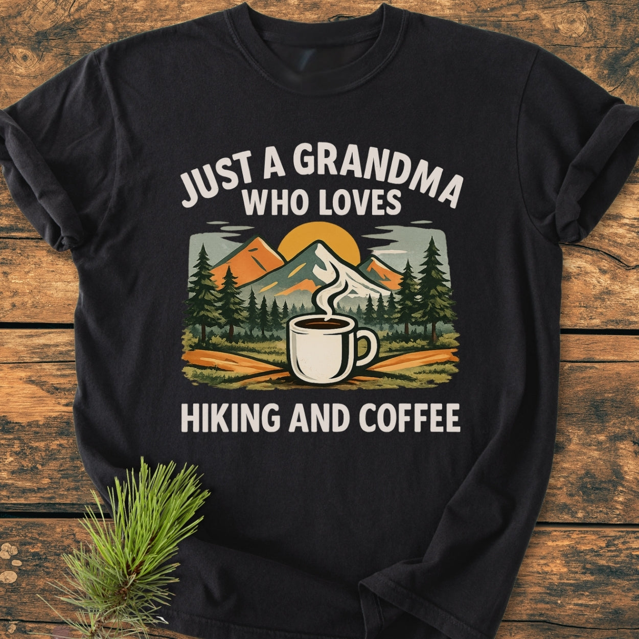 Grandma Loves Hiking And Coffee - Vintage Wash Tee features a colorful graphic of pine trees, mountains, and a coffee mug with hiking-themed text, laid out on a wooden surface.