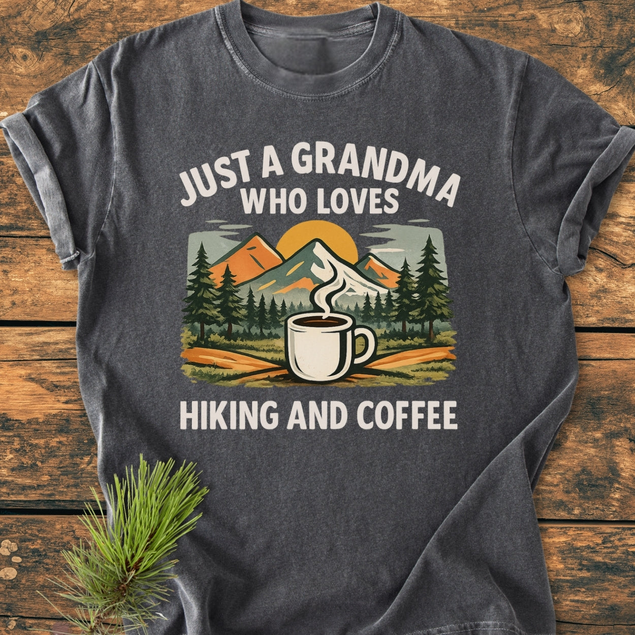 Grandma Loves Hiking And Coffee - Vintage Wash Tee features a stylized outdoor scene with pine trees, mountains, and a coffee mug, highlighting the text JUST A GRANDMA WHO LOVES HIKING AND COFFEE.