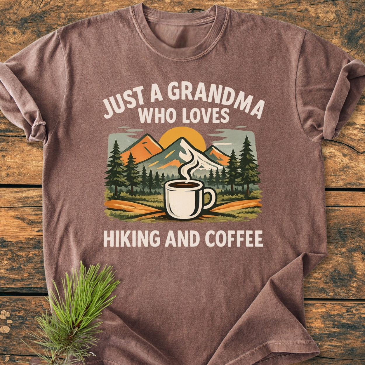 Grandma Loves Hiking And Coffee - Vintage Wash Tee featuring a colorful graphic of pine trees, mountains, and a coffee mug, laid on wood with a pine sprig, embodying comfort and durability.