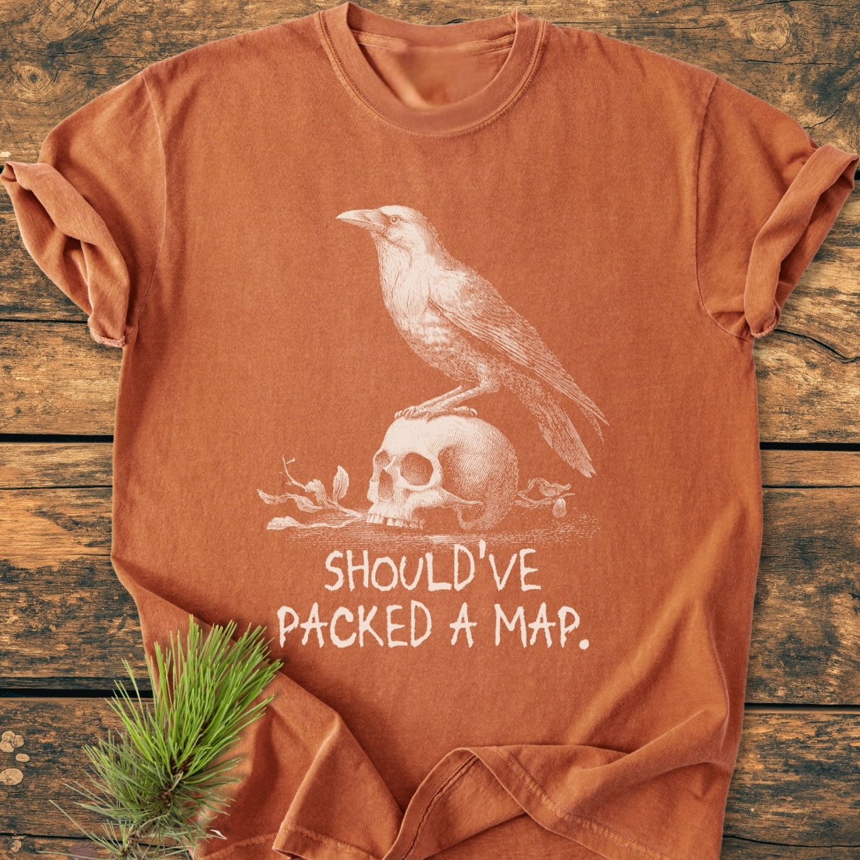 No WI-FI - Vintage Wash Tee: Burnt-orange shirt with crow and skull illustration, slogan “SHOULD’VE PACKED A MAP,” on wooden surface, styled with a pine sprig.
