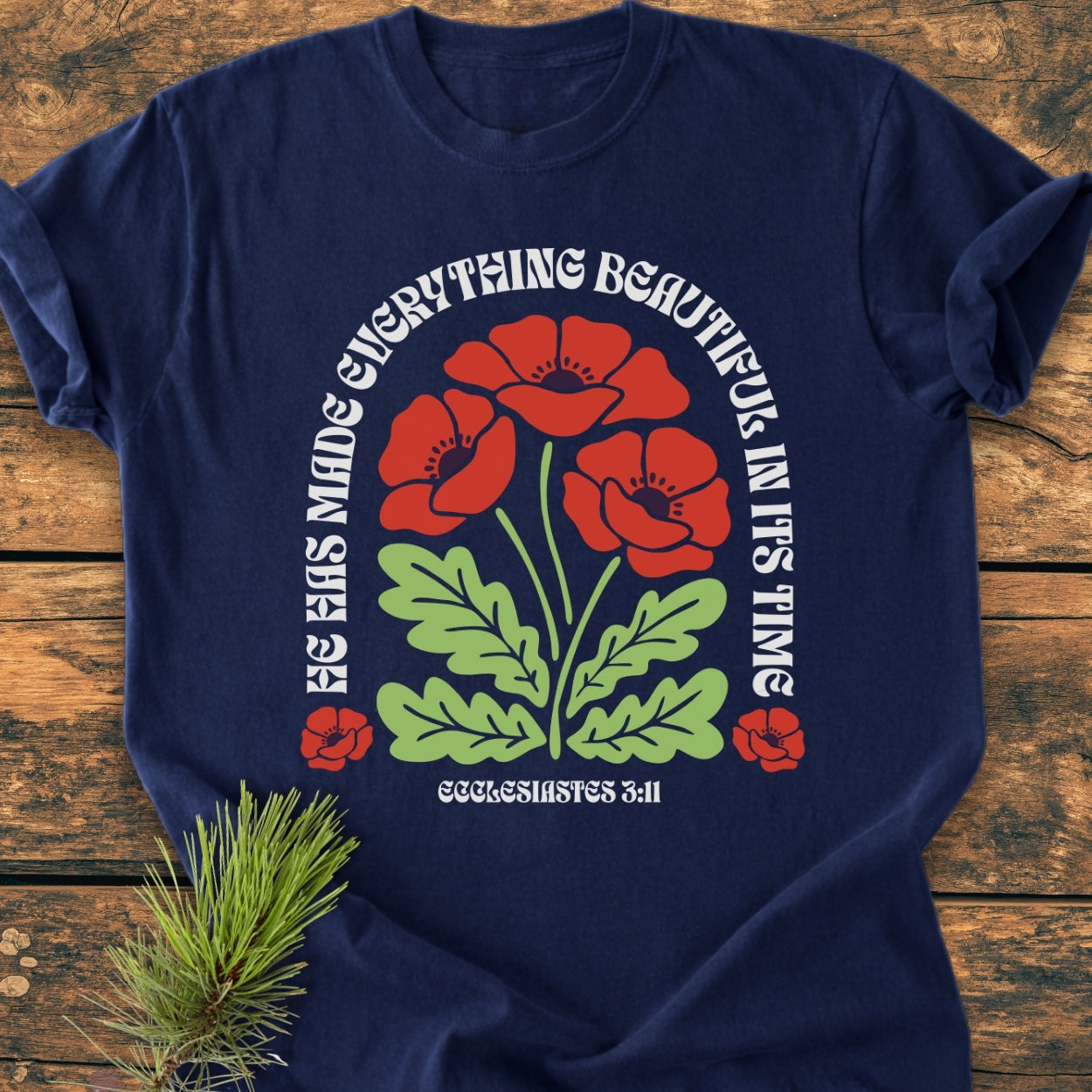 He Has Made Everything Beautiful - Vintage Wash Tee features a floral graphic with Ecclesiastes 3:11 on a navy-blue T-shirt, laid flat on wood with a pine sprig nearby.