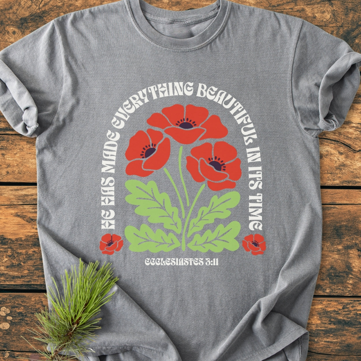 He Has Made Everything Beautiful - Vintage Wash Tee with stylized red poppies and Ecclesiastes 3:11 text on heather-gray fabric, laid on a wooden surface with a pine sprig.