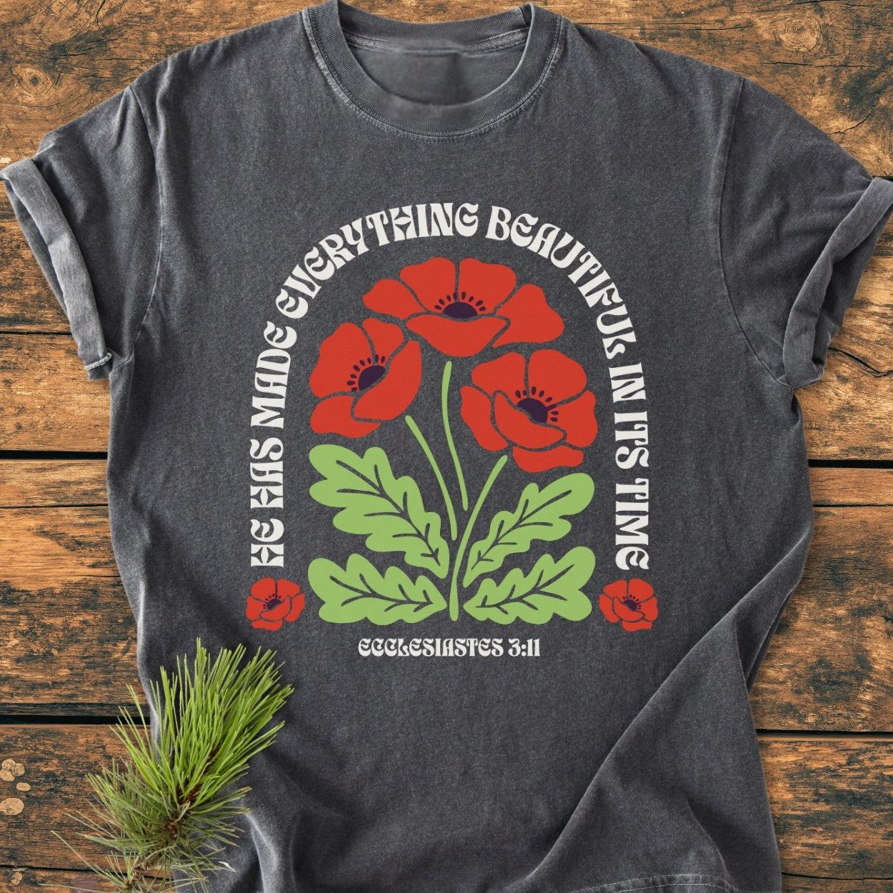 He Has Made Everything Beautiful - Vintage Wash Tee features a charcoal-gray color with red poppies and Bible verse from Ecclesiastes 3:11, laid flat on a wooden surface, showcasing its relaxed fit.