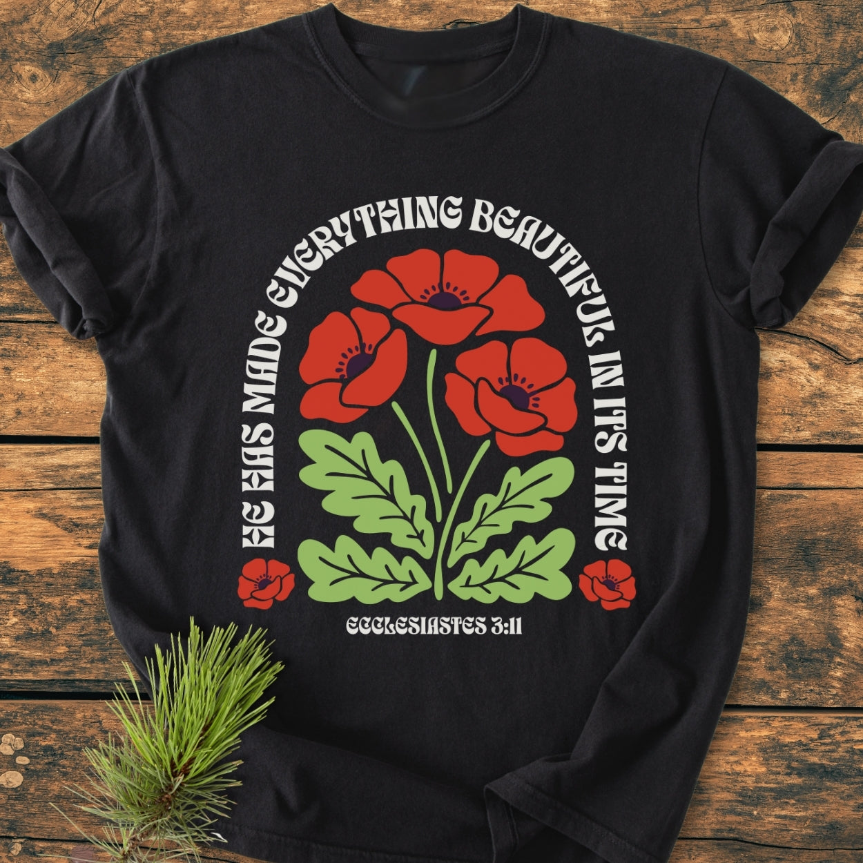 He Has Made Everything Beautiful - Vintage Wash Tee displays a stylized graphic of poppy flowers with Ecclesiastes 3:11 text on a black T-shirt, laid on a wooden surface with a pine sprig beside it.