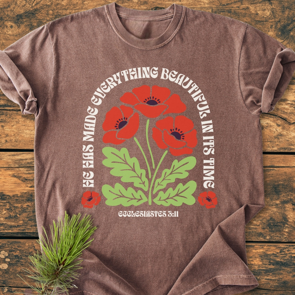 He Has Made Everything Beautiful - Vintage Wash Tee featuring red poppy-like flowers and Ecclesiastes 3:11 quote, on a brown short-sleeved T-shirt laid on a wooden surface.