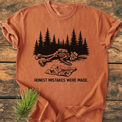 Honest Mistakes Were Made - Vintage Wash Tee featuring a cartoonish skeleton hiker scene, printed on a soft, durable garment-dyed T-shirt, displayed on a wooden surface with a pine sprig.