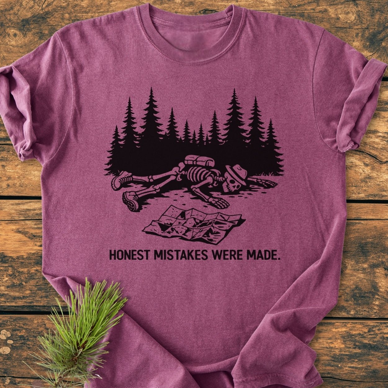 Honest Mistakes Were Made - Vintage Wash Tee features a cartoon skeleton hiking scene with a humorous caption, on a soft, relaxed fit, mauve garment-dyed T-shirt laid on a wooden surface.