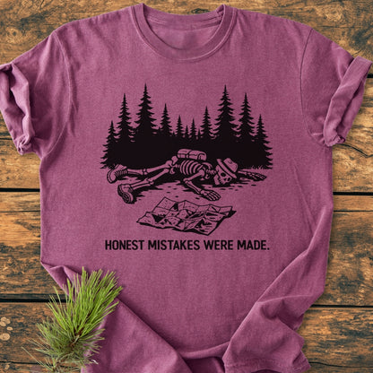 Honest Mistakes Were Made - Vintage Wash Tee features a cartoon skeleton hiking scene with a humorous caption, on a soft, relaxed fit, mauve garment-dyed T-shirt laid on a wooden surface.