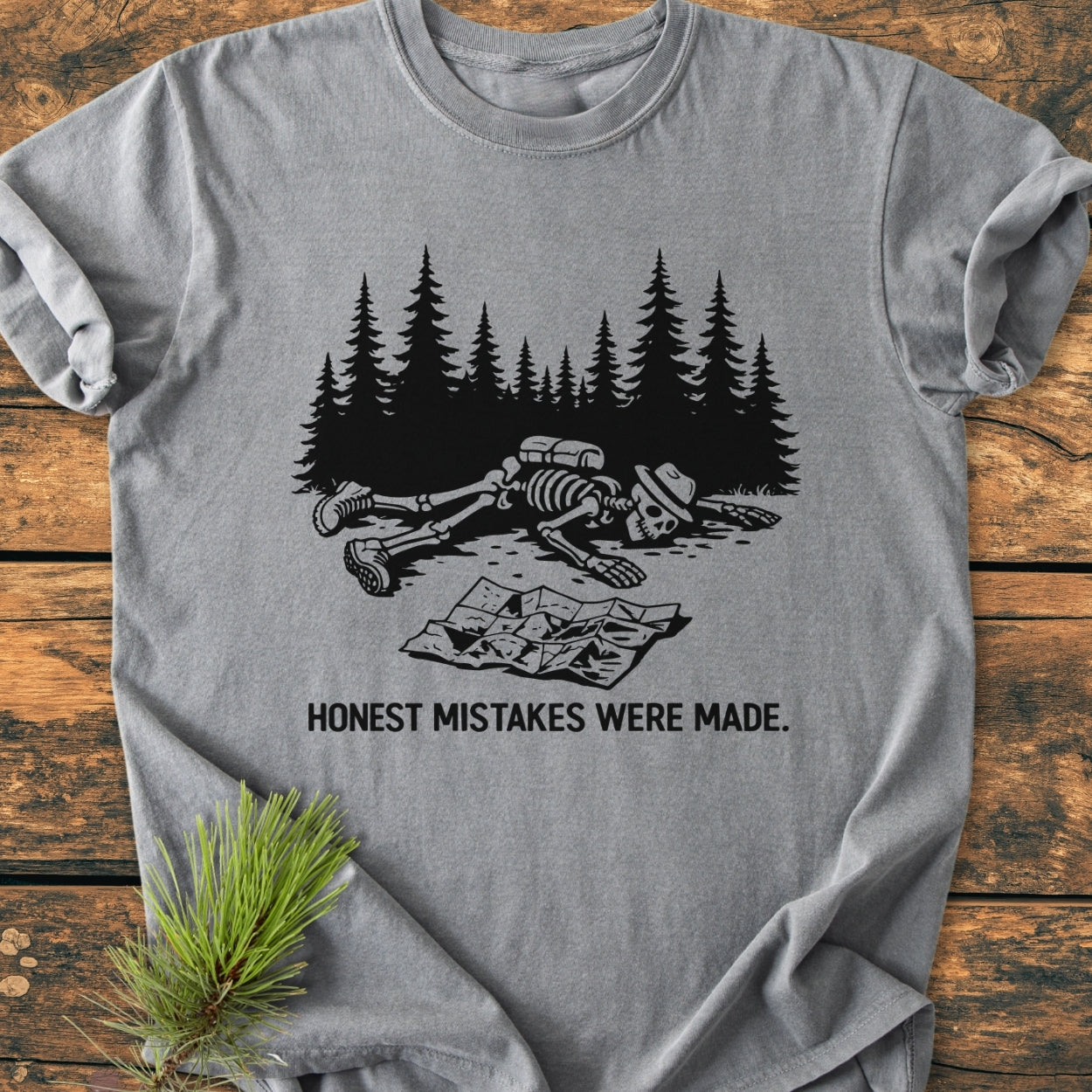 Heather-gray Honest Mistakes Were Made - Vintage Wash Tee featuring a black graphic of a collapsed hiker’s skeleton in front of pine trees with the caption, “HONEST MISTAKES WERE MADE,” laid flat with pine sprig.