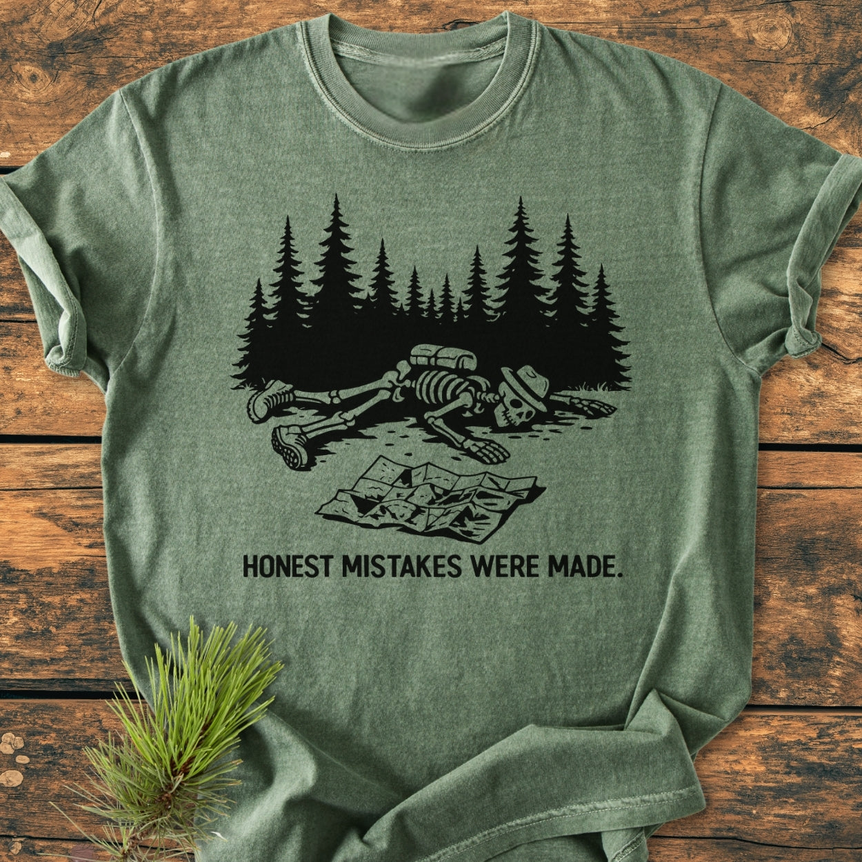 Honest Mistakes Were Made - Vintage Wash Tee features a black graphic of a hiker’s skeleton with gear, collapsed before pine trees, on a soft, heather-green, garment-dyed, and durable t-shirt.