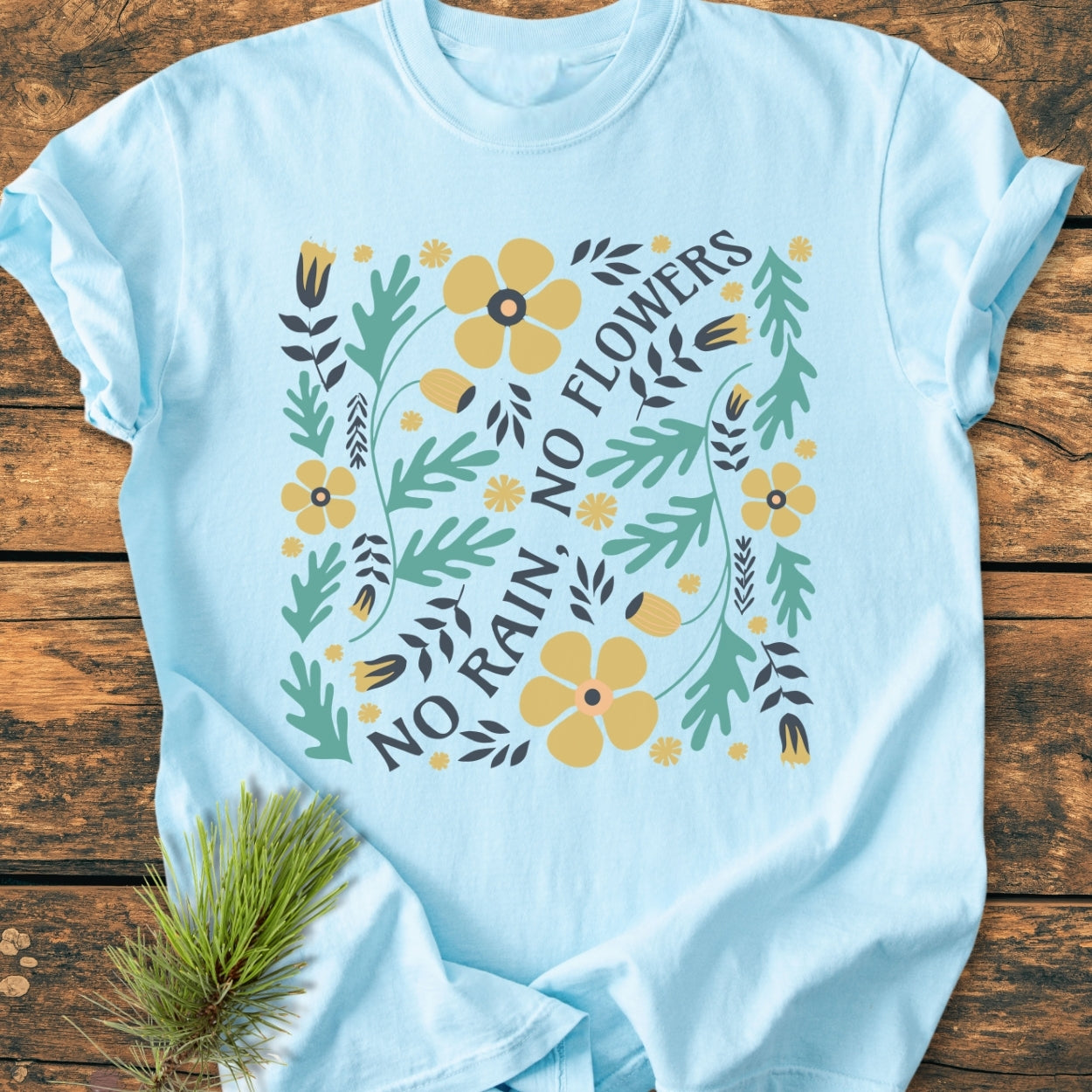 No Rain No Flowers - Vintage Wash Tee featuring a light-blue, soft-washed cotton fabric with a floral graphic and diagonal text, laid on wood with a pine sprig beside it.